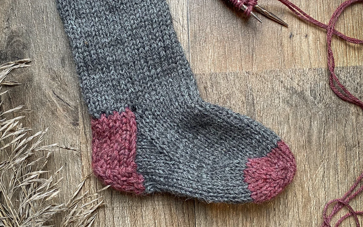 Handgestrickte graue Socke mit rosa Ferse auf Holzuntergrund.