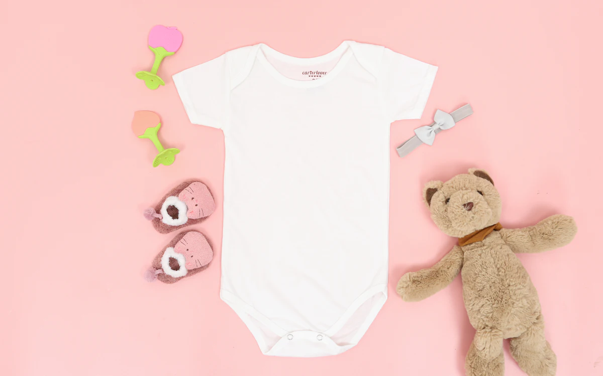Weißer Babybody flach ausgelegt auf rosa Hintergrund, daneben Teddybär und Babyschuhe.