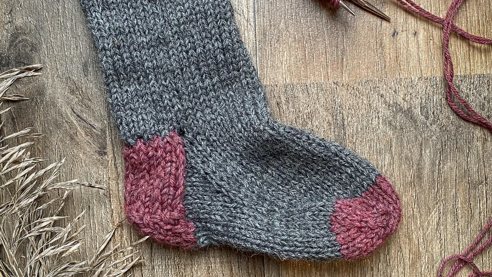 Handgestrickte graue Socke mit rosa Ferse und Spitze auf Holzuntergrund.